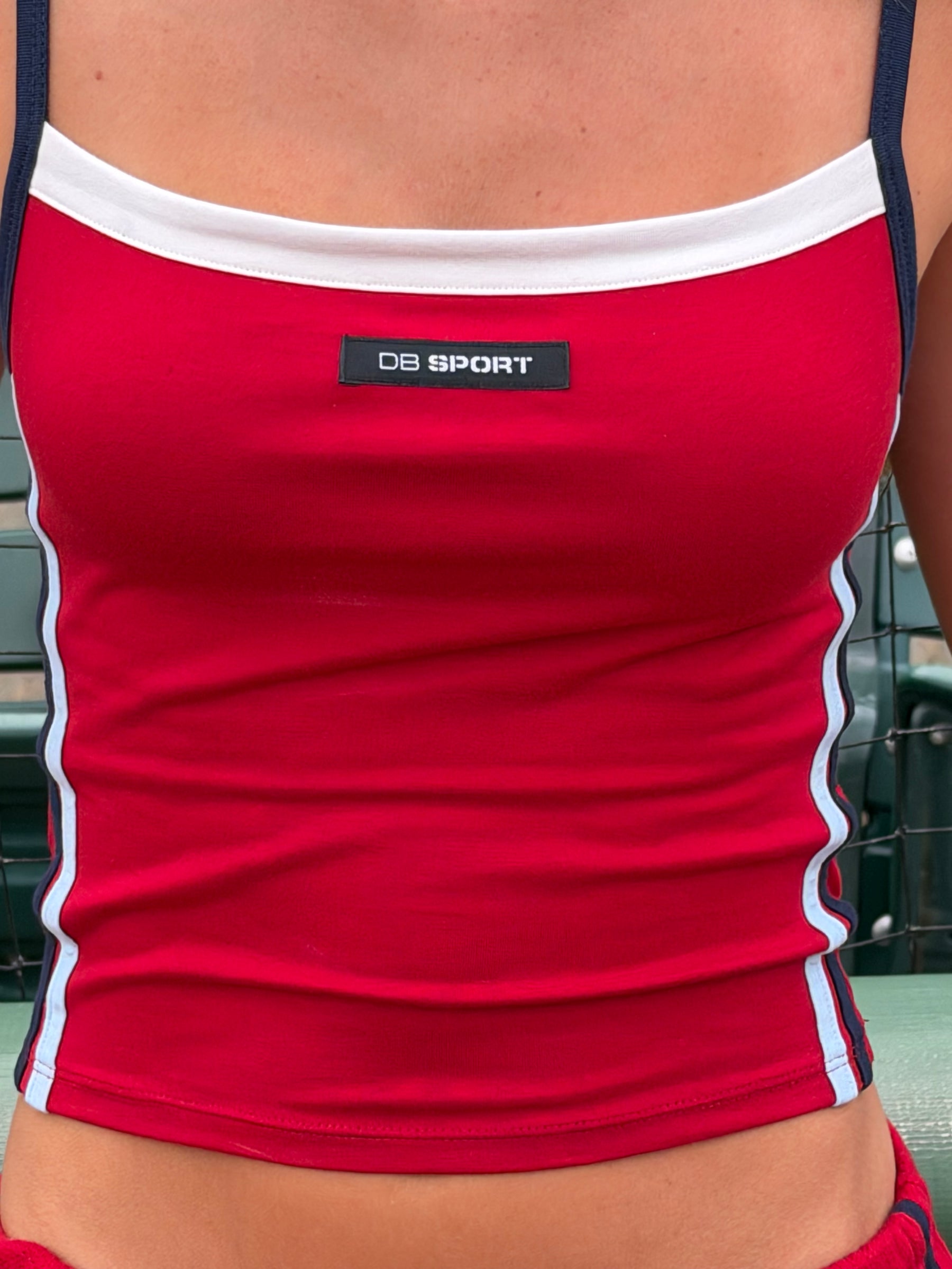A close-up of a person wearing a red athletic tank top with white trim and navy straps. A small black chest label reads 'DB SPORT'.
|M2