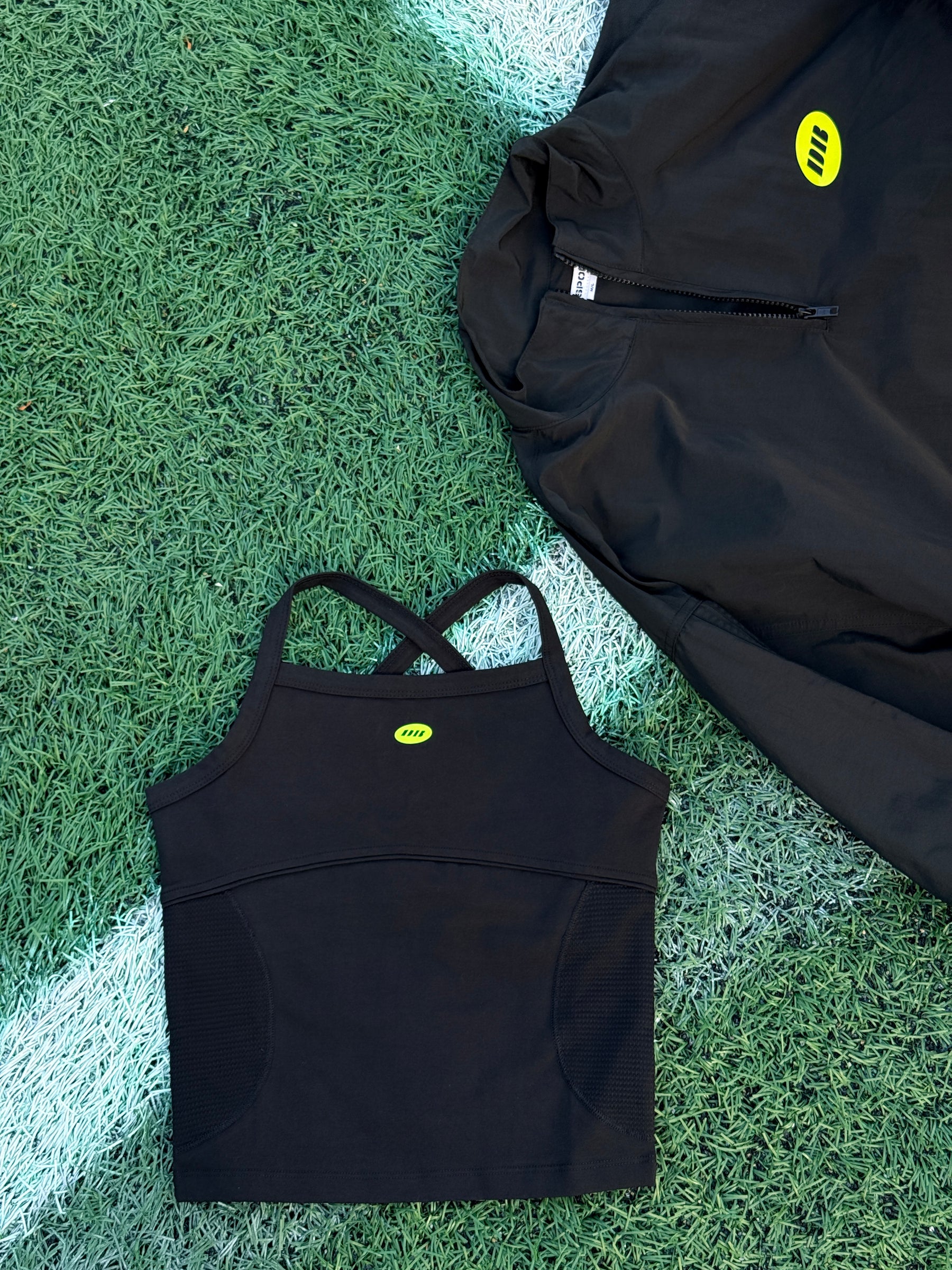 A black athletic zip-up jacket and a matching black sports bra laid flat on green turf with a white line, both featuring a small yellow logo.