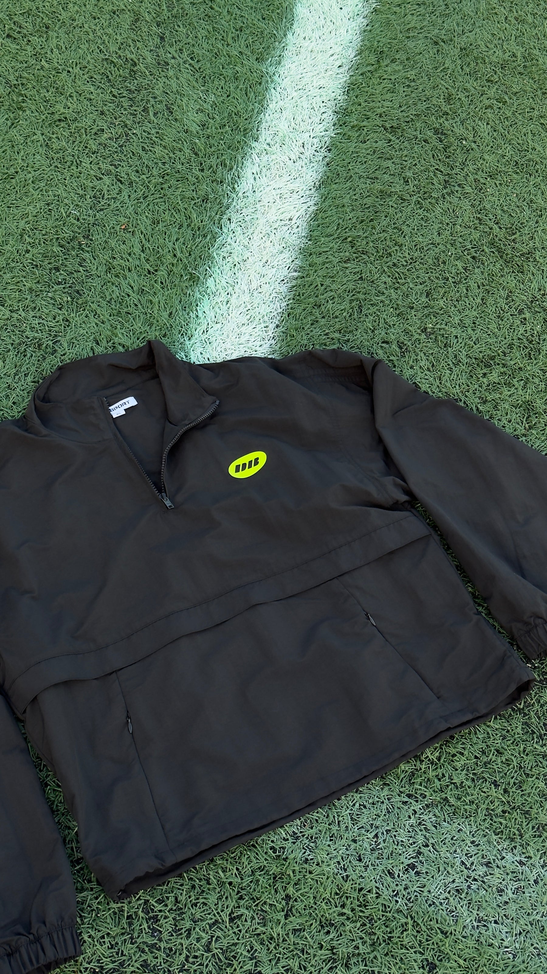 A black half-zip pullover jacket with a small yellow oval logo on the left chest, laid flat on green turf with a white boundary line.
