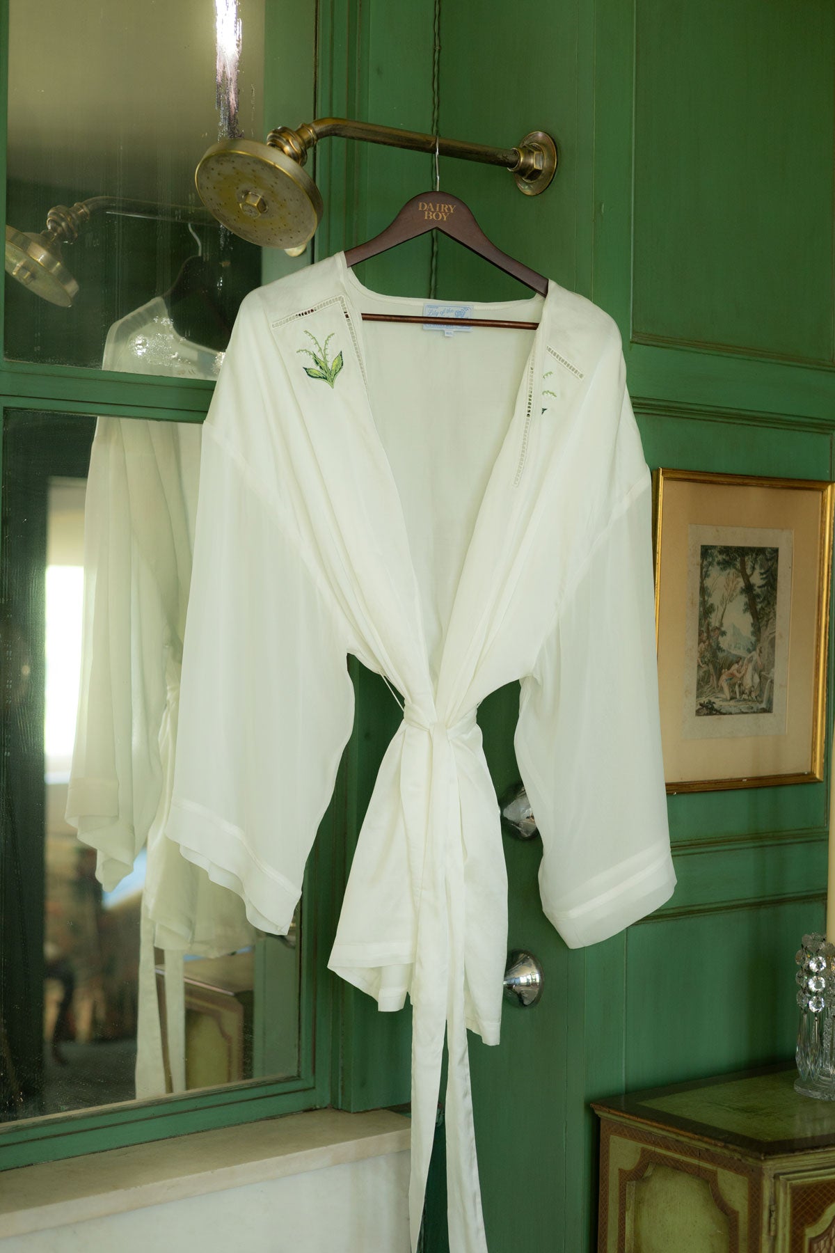 A white long-sleeve wrap robe with a tie waist and small green embroidered details on the chest, hanging on a wooden hanger against a green paneled wall.