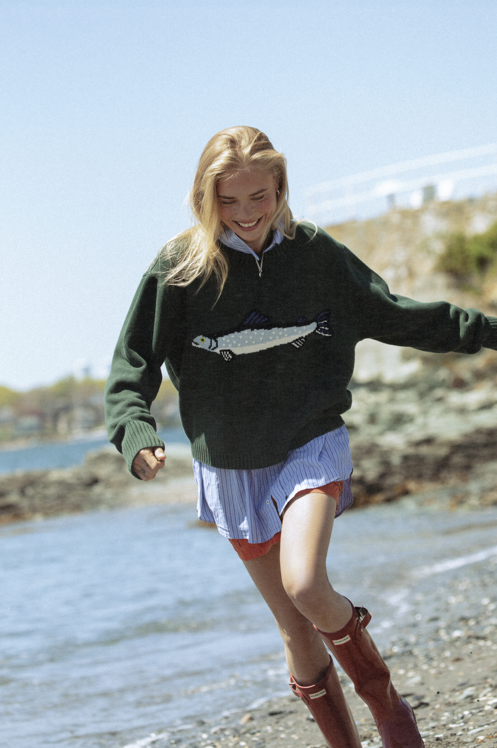 Woman wearing a green sweater with a fish design by a body of water.