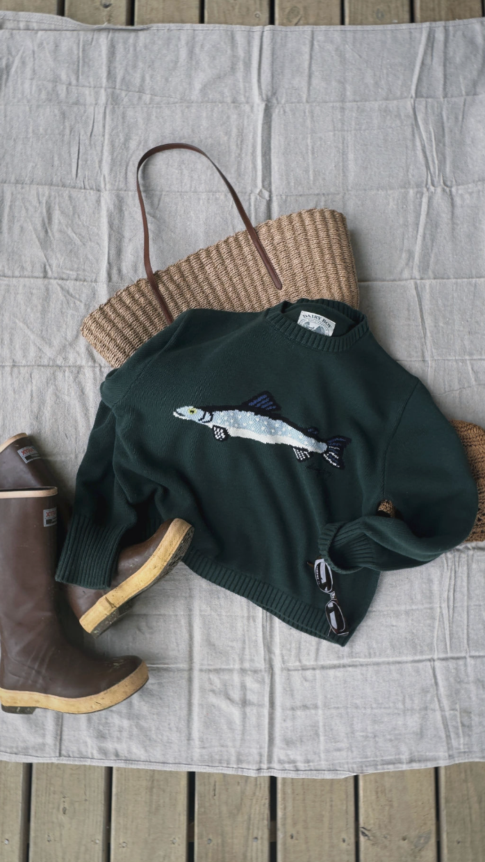 Green sweater with fish design, brown boots, and woven bag on a textured surface