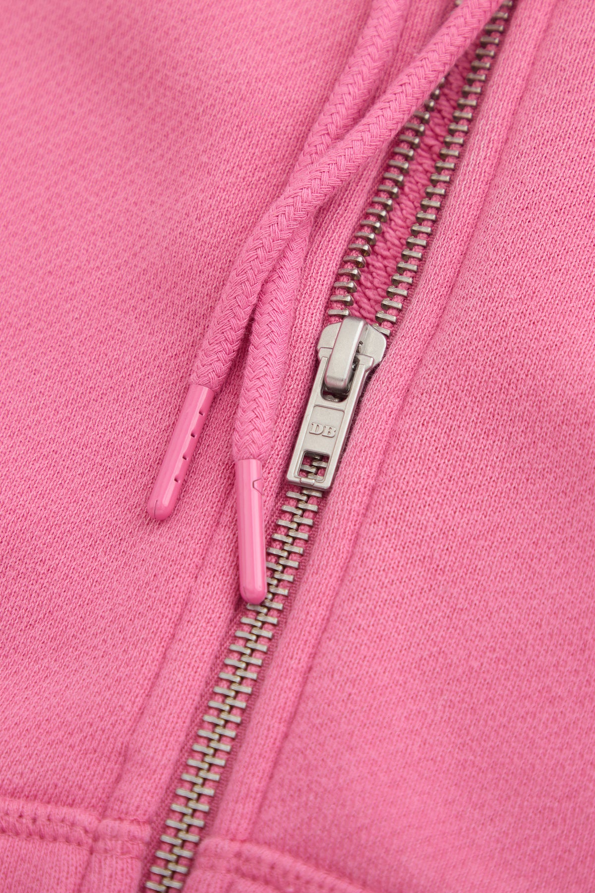 Close-up of the Dairy Boy Strawberry Milk Bottle Zip Up in strawberry pink, highlighting its metal zipper with DB engraved and two matching drawstrings with plastic tips, resting on the soft fabric.
