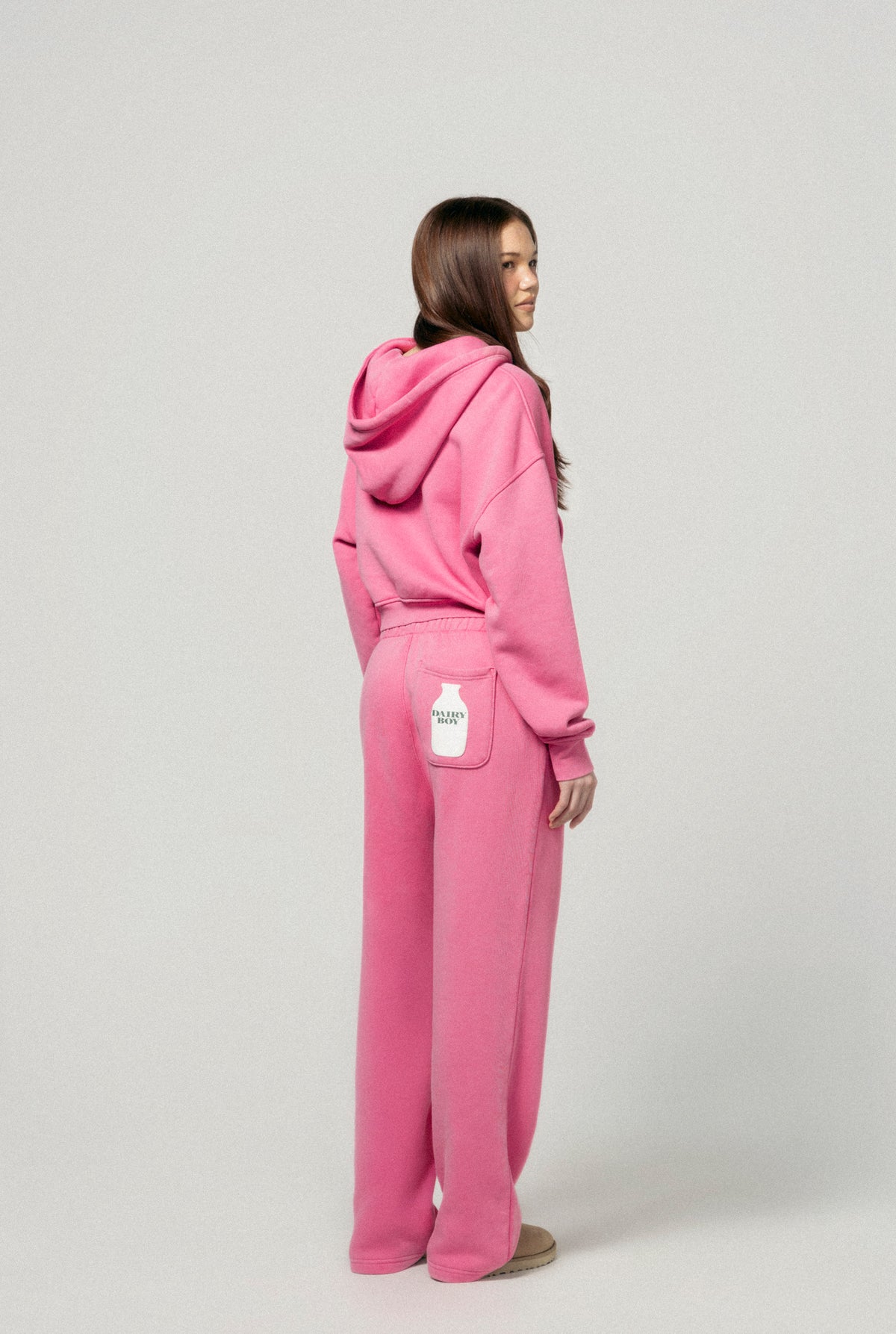 A woman faces away, wearing the Dairy Boy Strawberry Milk Bottle Zip Up and matching strawberry pink sweatpants with a white milk bottle on the right back pocket. The background is plain light gray. |m1