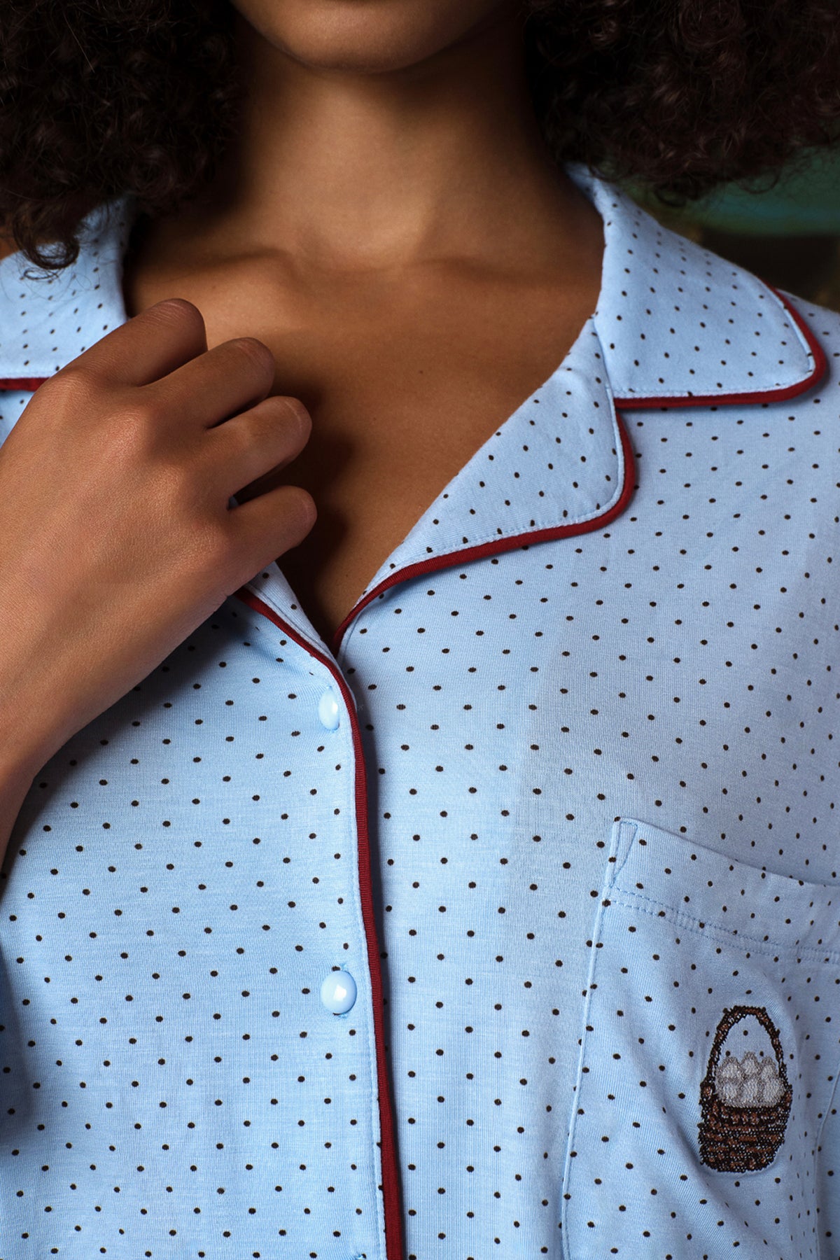 Close-up of a light blue pajama shirt with small dark polka dots, burgundy piping along the collar and button placket, and a chest pocket with a small basket motif, worn on a model.