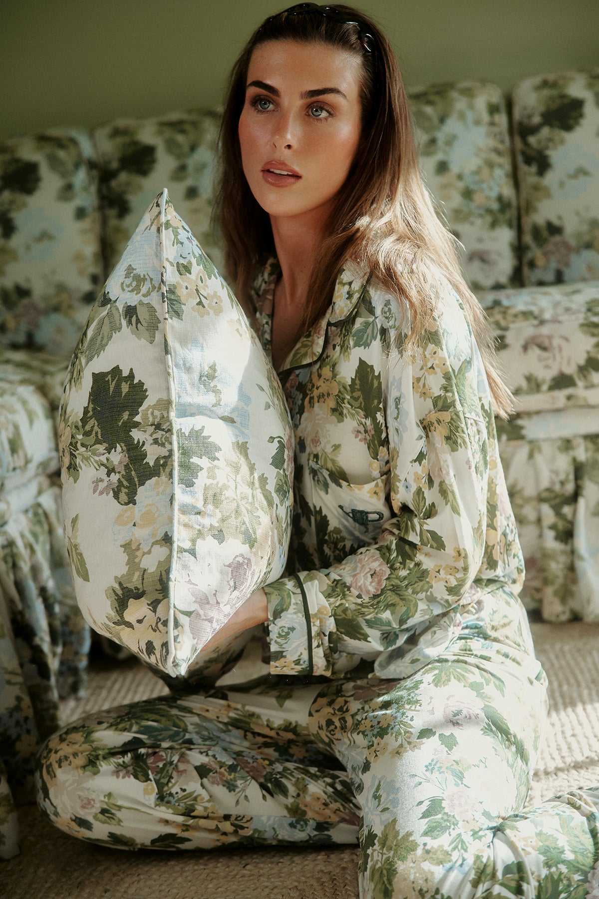 Long-sleeve pajama set in a green, cream and pale blue floral print with dark piping and a chest pocket, worn on a model holding a matching cushion. |M1