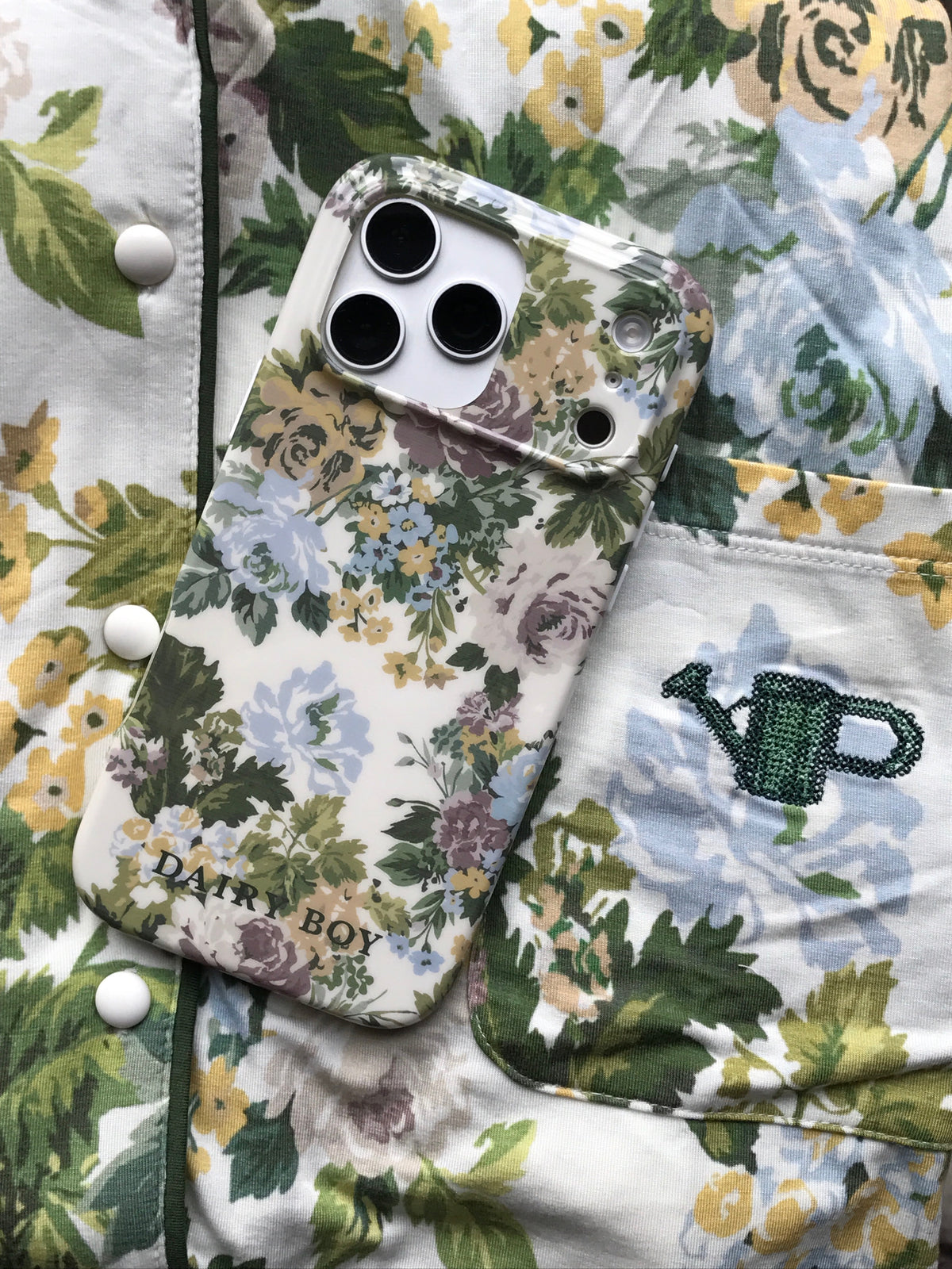 Phone case with a cream, blue, purple, yellow and green floral pattern and "DAIRY BOY" lettering, back view on floral pyjamas.