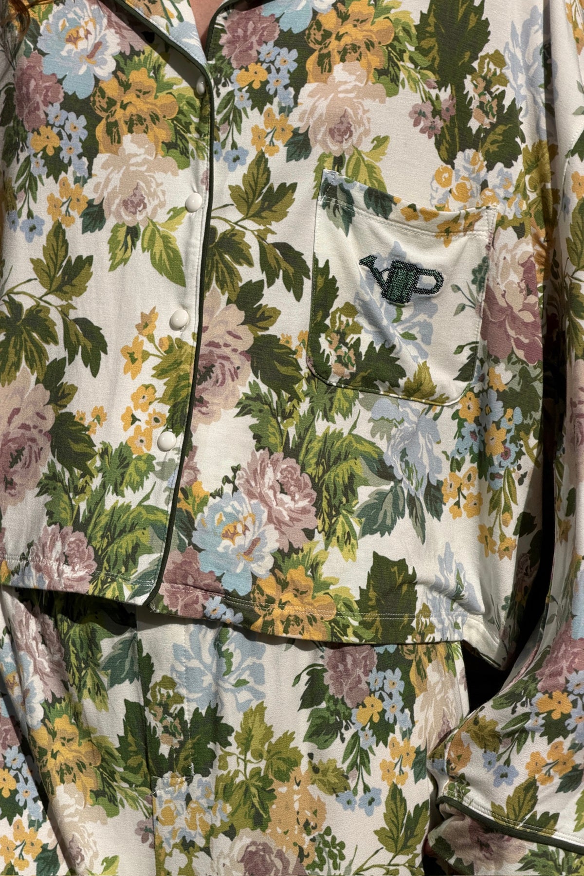 Close-up of a floral pajama shirt and pants set in cream with green, mauve, blue and yellow flowers, dark piping, snap buttons and a chest pocket with an embroidered watering can.