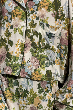 Close-up of a floral pajama shirt and pants set in cream with green, mauve, blue and yellow flowers, dark piping, snap buttons and a chest pocket with an embroidered watering can.