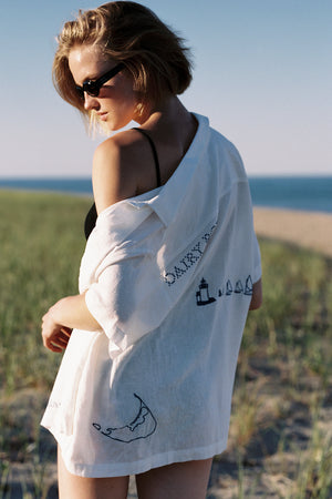 Woman wearing a white shirt with text and graphics, standing on a beach.