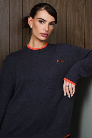 A woman wearing a dark grey crewneck sweater with bright orange trim at the collar and cuffs, featuring the embroidered initials 'D.B.
|M1