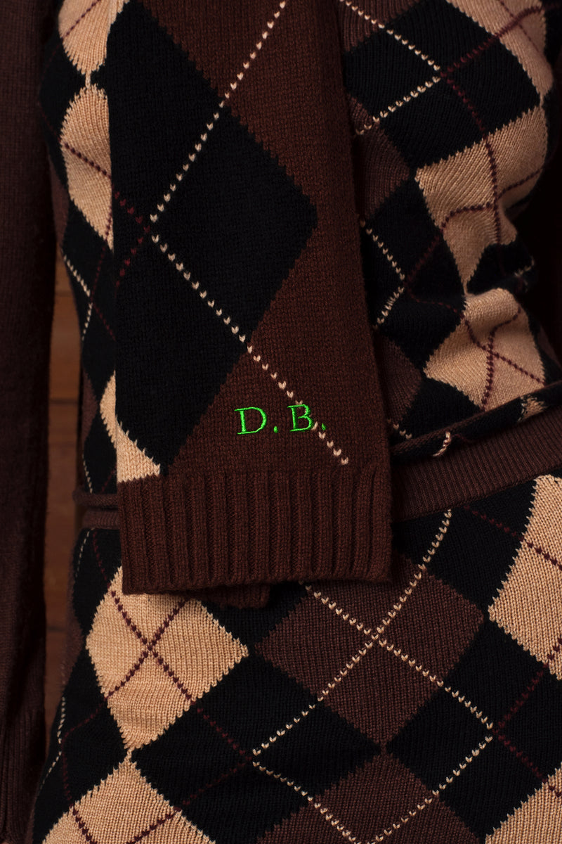Close-up of a knitted scarf with an argyle pattern in dark brown, black, and beige, featuring a ribbed trim and green embroidered letters 'D. B.
