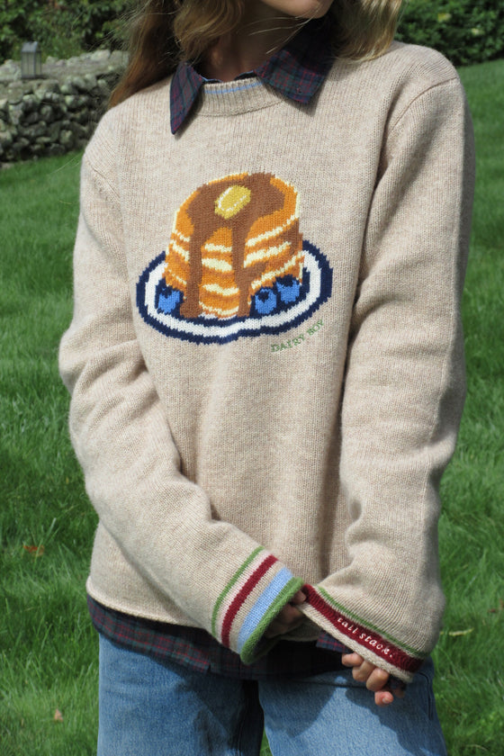 A person wearing a cream knit sweater with a knitted graphic of pancakes on a plate, layered over a plaid shirt and blue jeans, standing outdoors on a grassy lawn.
|M1