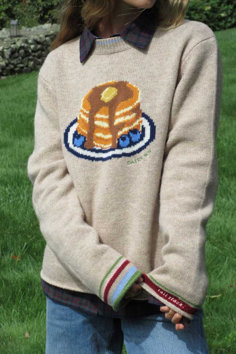 A person wearing a cream knit sweater with a knitted graphic of pancakes on a plate, layered over a plaid shirt and blue jeans, standing outdoors on a grassy lawn.
|M1