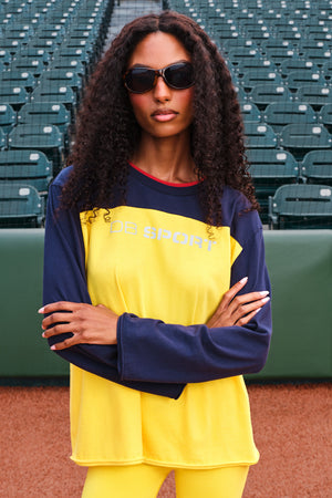 A woman with curly hair and sunglasses stands on a baseball field wearing a two-tone athletic long-sleeve shirt and matching yellow pants. The shirt is bright yellow on the front with navy sleeves and collar, featuring 'DB SPORT' printed across the chest.
|M1