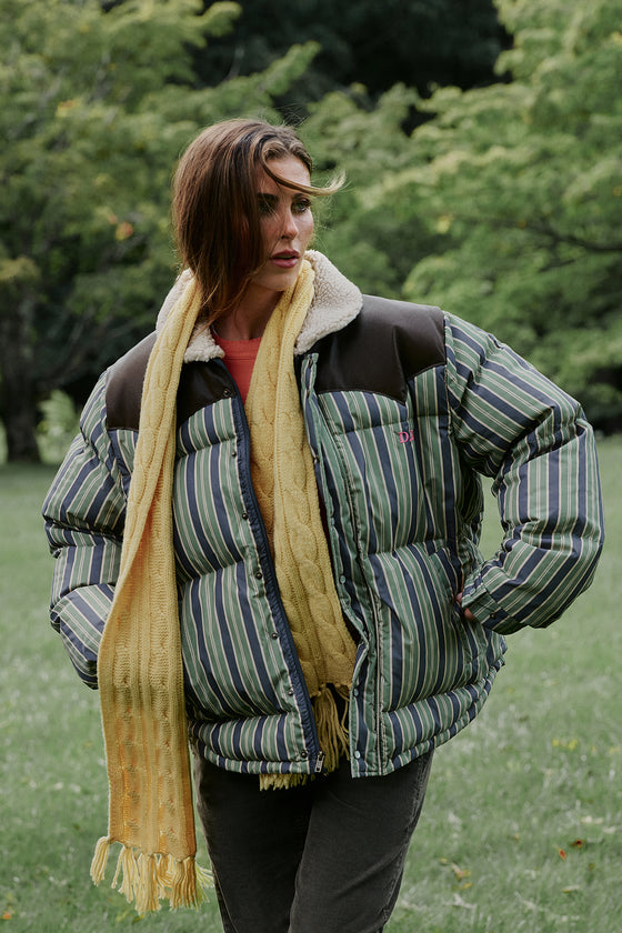 A woman standing outdoors in a grassy park wearing a puffer jacket with green and blue vertical stripes, dark brown shoulder panels, and a cream sherpa collar, styled with a chunky yellow cable-knit scarf.
|M2