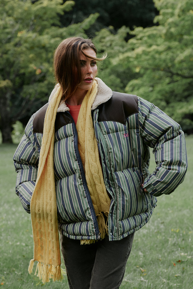 A woman standing outdoors in a grassy park wearing a puffer jacket with green and blue vertical stripes, dark brown shoulder panels, and a cream sherpa collar, styled with a chunky yellow cable-knit scarf.
|M2