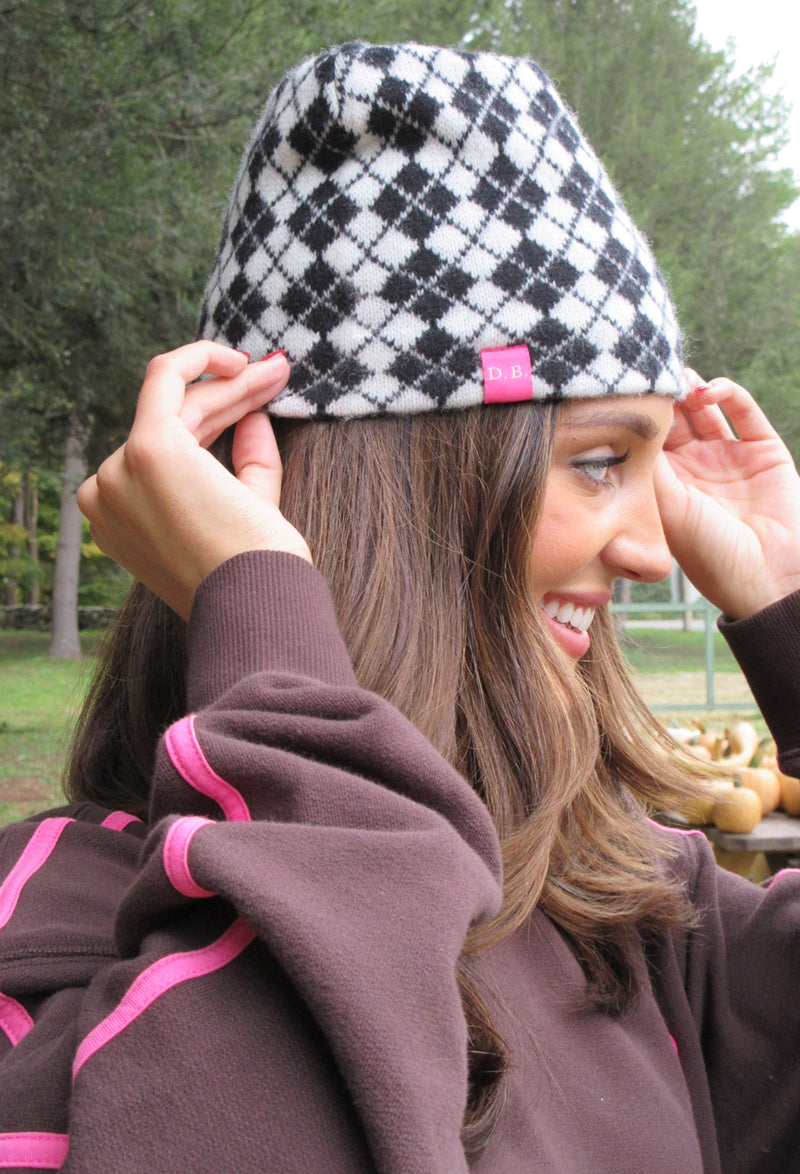 A woman outdoors wearing a black and white argyle knit beanie with a small pink tag, adjusting the brim with both hands. She is wearing a brown sweatshirt with pink stripes.