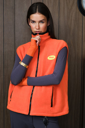 A woman wearing a bright orange fleece vest over a grey long-sleeve top, standing against a dark wood-paneled wall. The vest has a yellow circular logo on the left chest and black zippers.
|M1