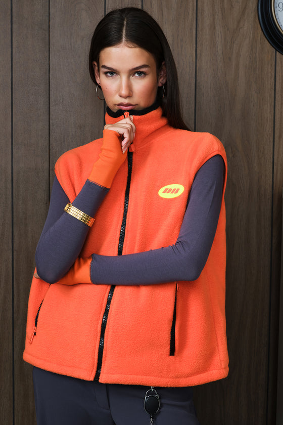 A woman wearing a bright orange fleece vest over a grey long-sleeve top, standing against a dark wood-paneled wall. The vest has a yellow circular logo on the left chest and black zippers.
|M1