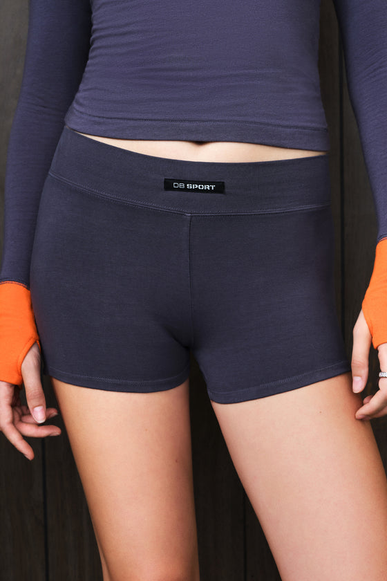 A close-up of a model wearing dark grey athletic shorts with a visible 'DB SPORT' brand label on the waistband, paired with a matching long-sleeve top featuring orange cuffs.
|M1