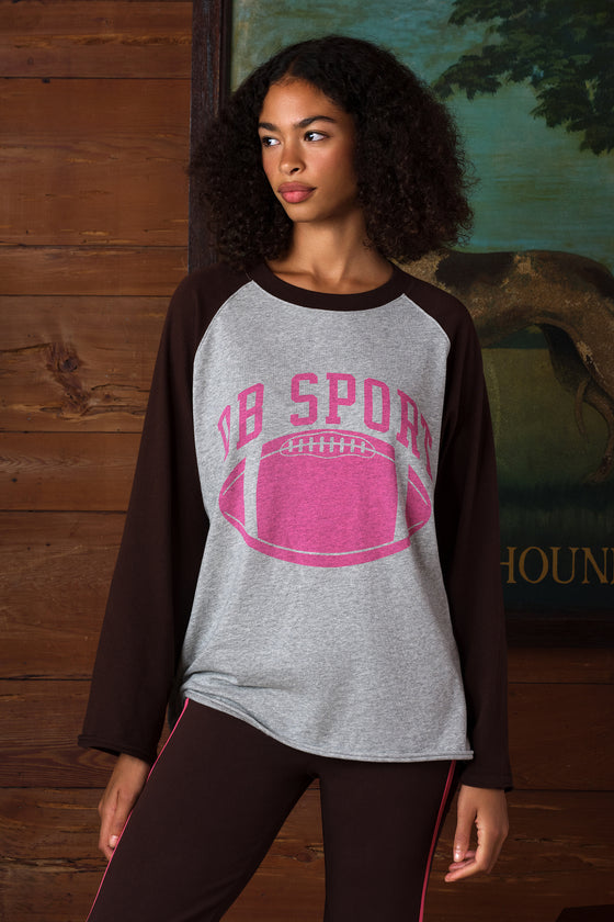 A woman wearing a heather gray and dark brown raglan long-sleeve t-shirt with a pink 'DB SPORT' football graphic on the front, posed against a rustic wood-paneled background.
|M1