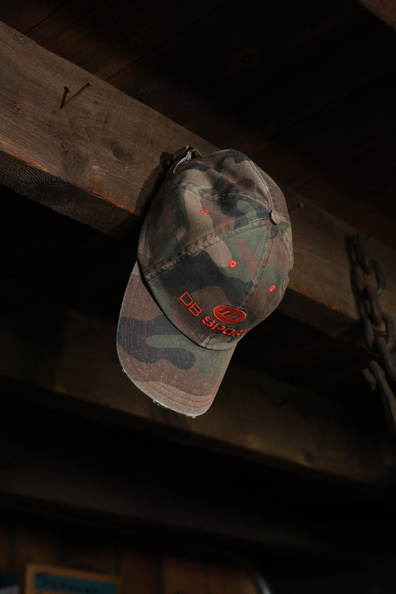 A camouflage baseball cap with red embroidered 'DB Sport' branding hanging from a rustic wooden beam in a dimly lit setting.