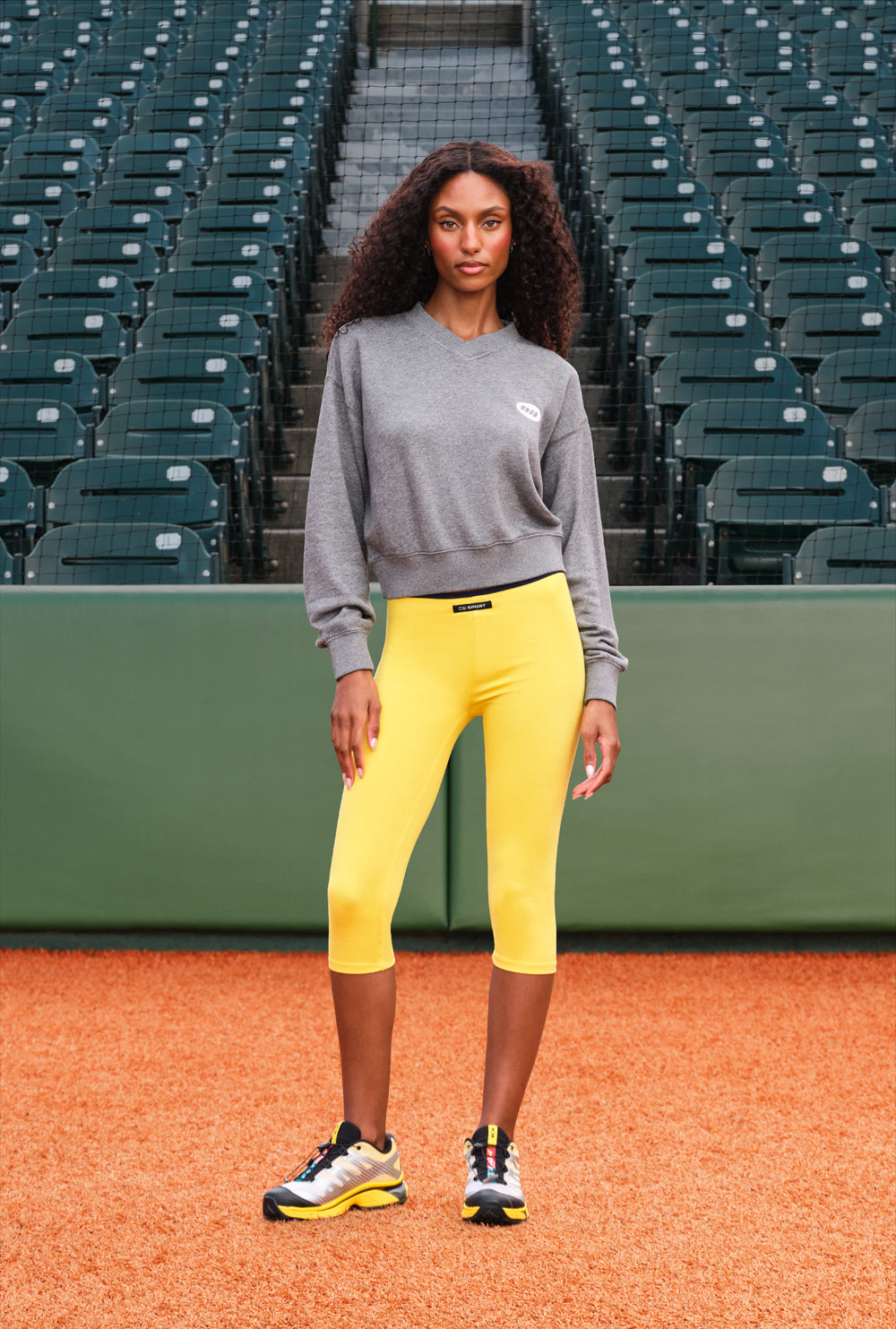 A woman stands on a baseball diamond wearing a gray sweatshirt, bright yellow capri leggings, and athletic sneakers, with rows of stadium seats in the background.
|M1