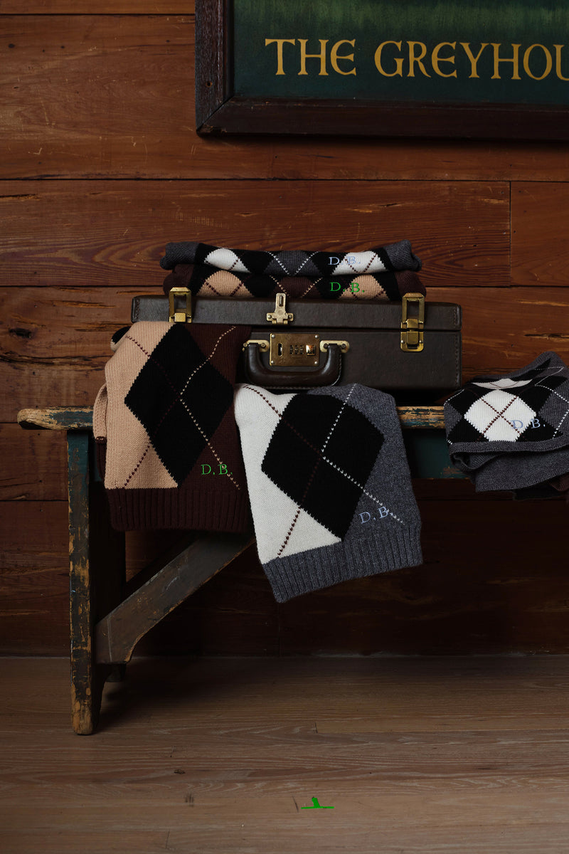 A styled product display of multiple pairs of argyle knit socks in various colors (black, white, brown, grey, beige) draped over and resting on a vintage dark brown suitcase, set against a rustic wooden wall and floor.
