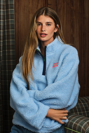 A woman wearing a light blue sherpa fleece quarter-zip pullover with a small pink embroidered logo on the chest, posed in front of a dark wood-paneled wall.
|M1