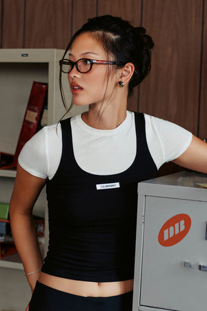A woman wearing a black  tank top over a white short-sleeve crewneck t-shirt, paired with black glasses and a black top, leaning against a gray metal cabinet in an indoor setting.
|M1