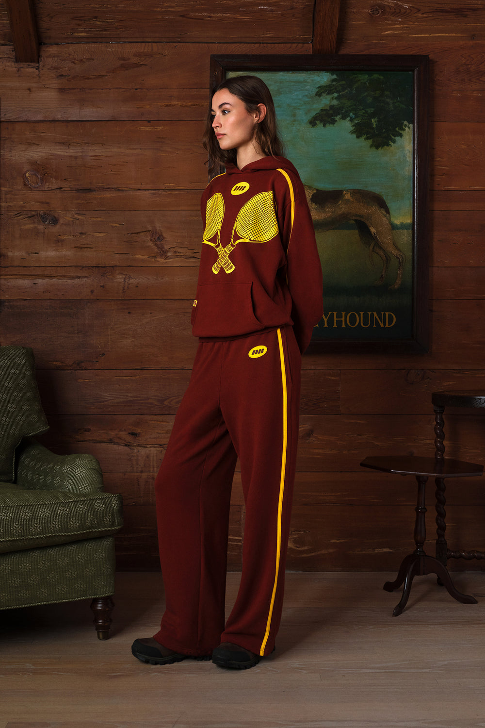 A woman stands in a rustic room with wood-paneled walls, wearing a burgundy sweatsuit with yellow accents. The set includes an oversized hoodie with a yellow tennis racket graphic on the chest and matching wide-leg pants with a yellow side stripe.
|M1