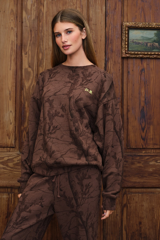 A woman modeling a matching brown camo patterned sweatshirt and sweatpants set against a rustic wooden paneled wall.
|M1
