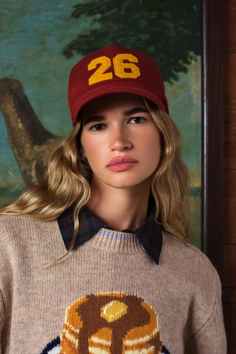 A woman wearing a maroon baseball cap with yellow '26' embroidery and a beige knit sweater, photographed against an indoor painted background.