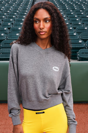 A woman with long curly dark hair stands in a stadium wearing a heather gray crewneck sweatshirt with a small white oval logo on the left chest, paired with bright yellow pants.
|M1