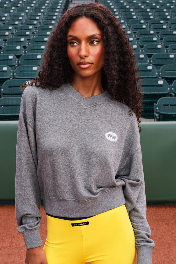 A woman with long curly dark hair stands in a stadium wearing a heather gray crewneck sweatshirt with a small white oval logo on the left chest, paired with bright yellow pants.
|M1