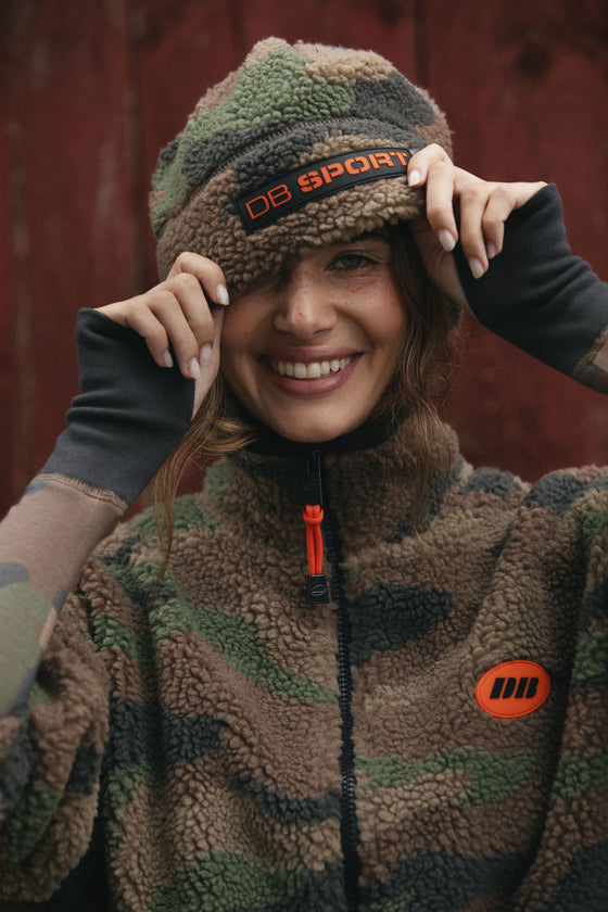 A smiling woman wearing a brown and green camouflage fleece jacket and a matching camouflage beanie with an orange 'DB SPORT' logo, posing in front of a rustic red-brown wooden background.