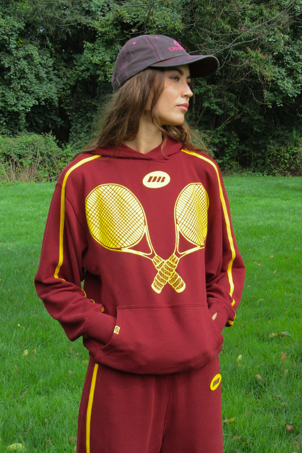 A woman wearing a maroon and yellow tennis-themed tracksuit with a large crossed-racquet graphic on the front, standing outdoors on a grassy lawn with trees in the background.
|M1