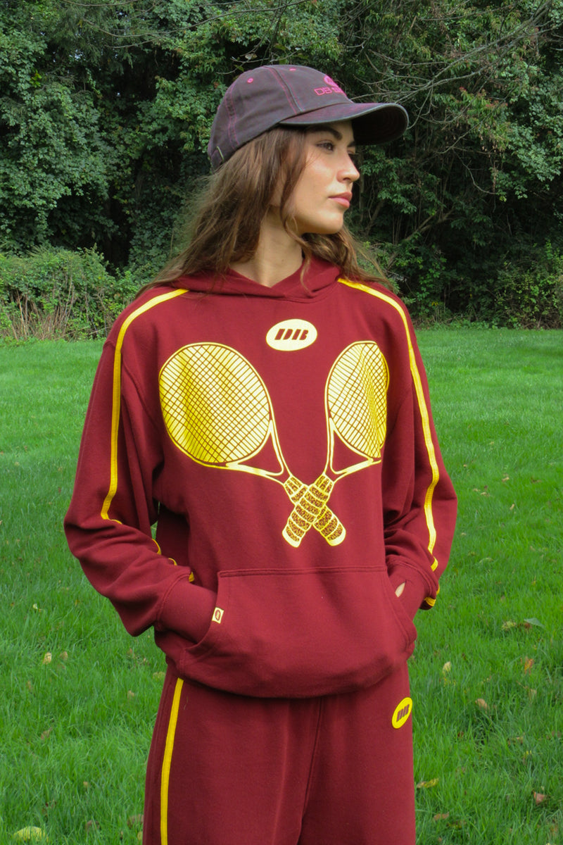 A woman wearing a maroon and yellow tennis-themed tracksuit with a large crossed-racquet graphic on the front, standing outdoors on a grassy lawn with trees in the background.
|M1