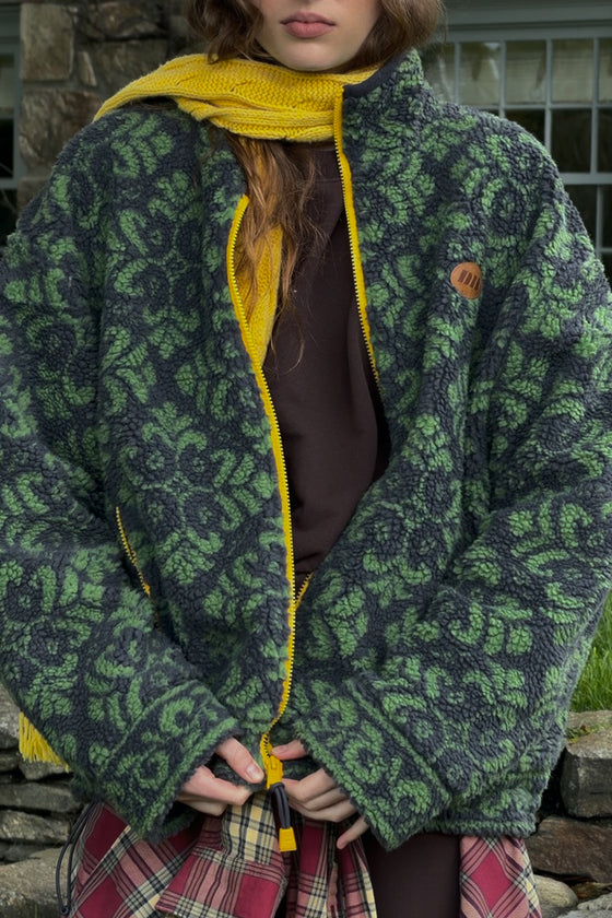 A person wearing a fuzzy zip-front jacket with a green and dark gray floral pattern, a bright yellow knit hood and zipper trim, and a brown top, photographed outdoors in front of a stone wall.
|M1