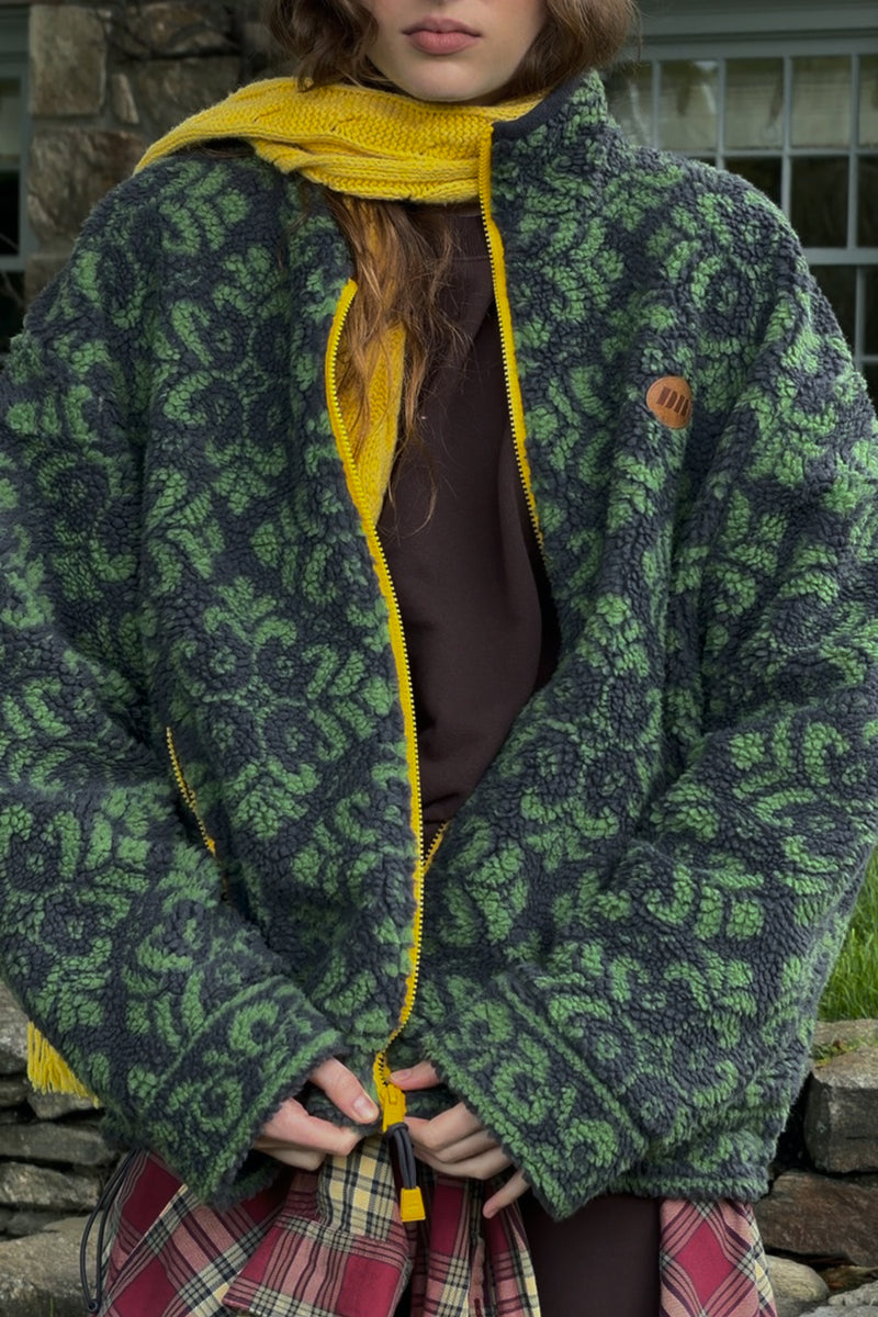 A person wearing a fuzzy zip-front jacket with a green and dark gray floral pattern, a bright yellow knit hood and zipper trim, and a brown top, photographed outdoors in front of a stone wall.
|M1