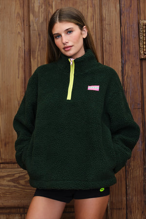 A woman wearing a dark forest green sherpa fleece pullover with a bright yellow zipper and a small pink logo patch on the chest, standing against a rustic wooden plank wall.
|M1