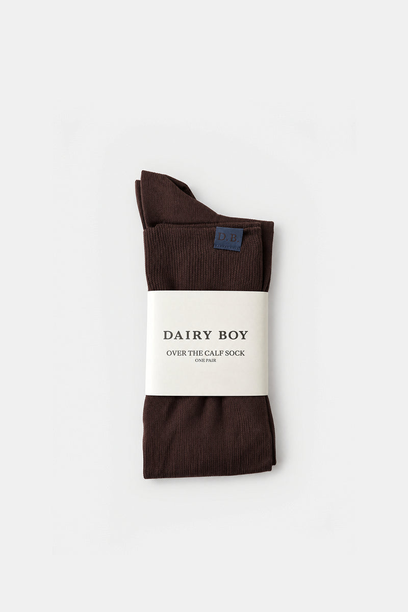 A pair of folded dark brown over-the-calf socks with a white paper label reading 'DAIRY BOY OVER THE CALF SOCK', photographed on a clean white background.