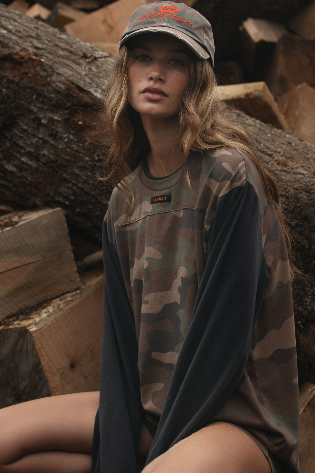 A woman wearing an oversized camouflage crewneck shirt with solid dark sleeves and a matching camouflage baseball cap, sitting among stacked logs.
|M1