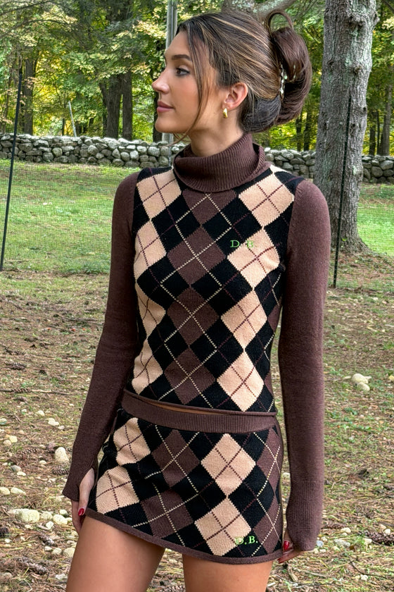 A woman wearing a brown and beige argyle turtleneck sweater and matching mini skirt, standing outdoors in a wooded area.
|M1
