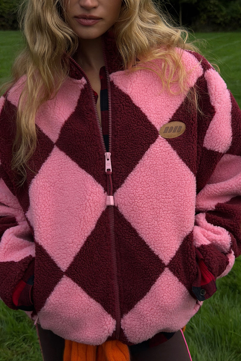 A close-up of a woman wearing a pink and burgundy argyle fleece zip-up jacket outdoors on grass. A small oval patch on the chest reads 'DB'.
|M1
