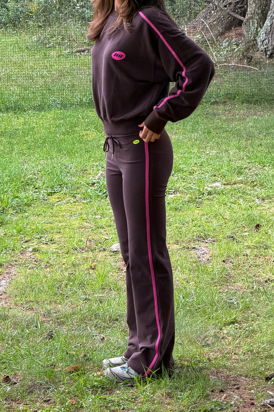 A person wearing a dark brown tracksuit with bright pink stripes and a pink chest logo, standing outdoors on a grassy lawn.
|M1