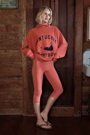 Person wearing a coral sweatshirt and leggings with 'Nantucket' text in a rustic setting.
|M1