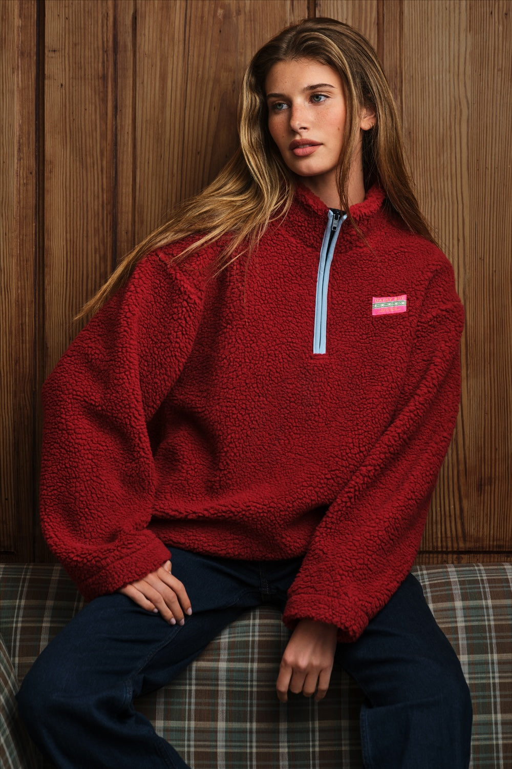 A woman wearing a red sherpa fleece quarter-zip pullover with a light blue zipper and a small pink chest logo, paired with dark blue denim, sitting in front of a rustic wooden wall.
|M1