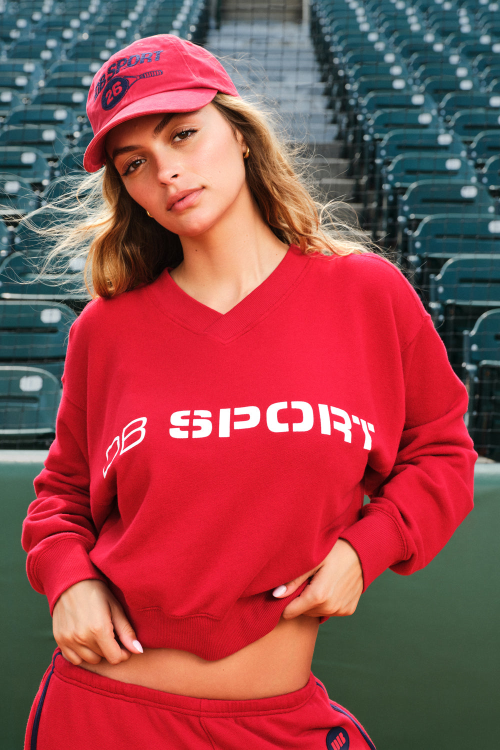 A woman wearing a red cropped V-neck sweatshirt with 'DB SPORT' printed in white across the chest, matching red sweatpants, and a red baseball cap, posed in front of stadium seating.
|M1